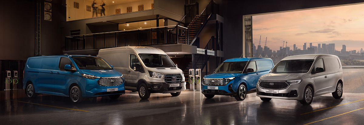 Vier Nutzfahrzeuge in einer Lagerhalle, darunter ein blauer Kastenwagen und ein grauer Transporter, mit Stadtansicht im Hintergrund.